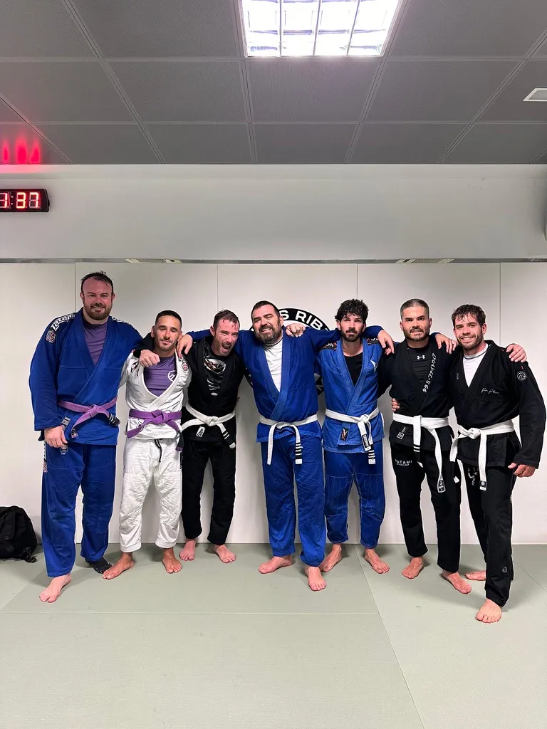 Siete practicantes de BJJ con cinturones blancos y morados posan juntos en el tatami de la academia.