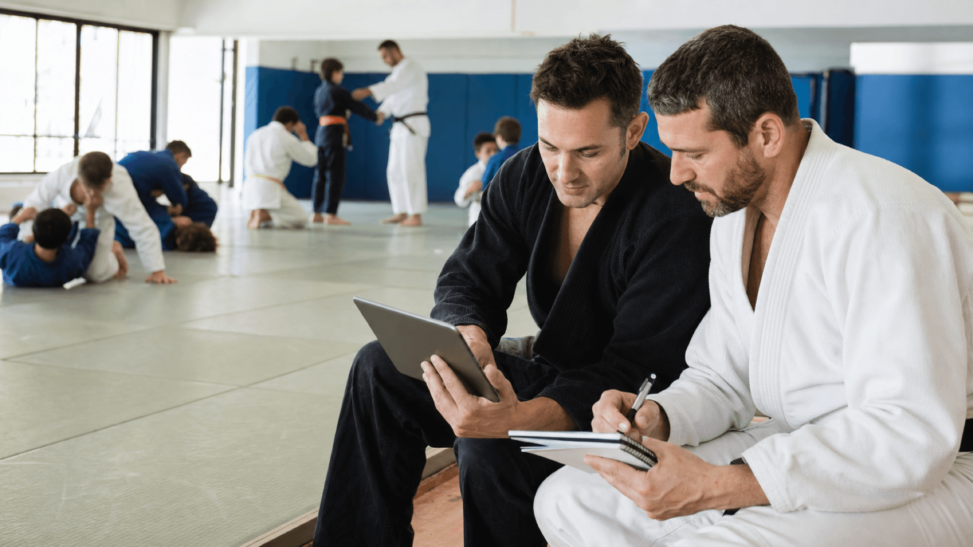 Dos instructores planifican junto al tatami mientras un grupo entrena en su academia